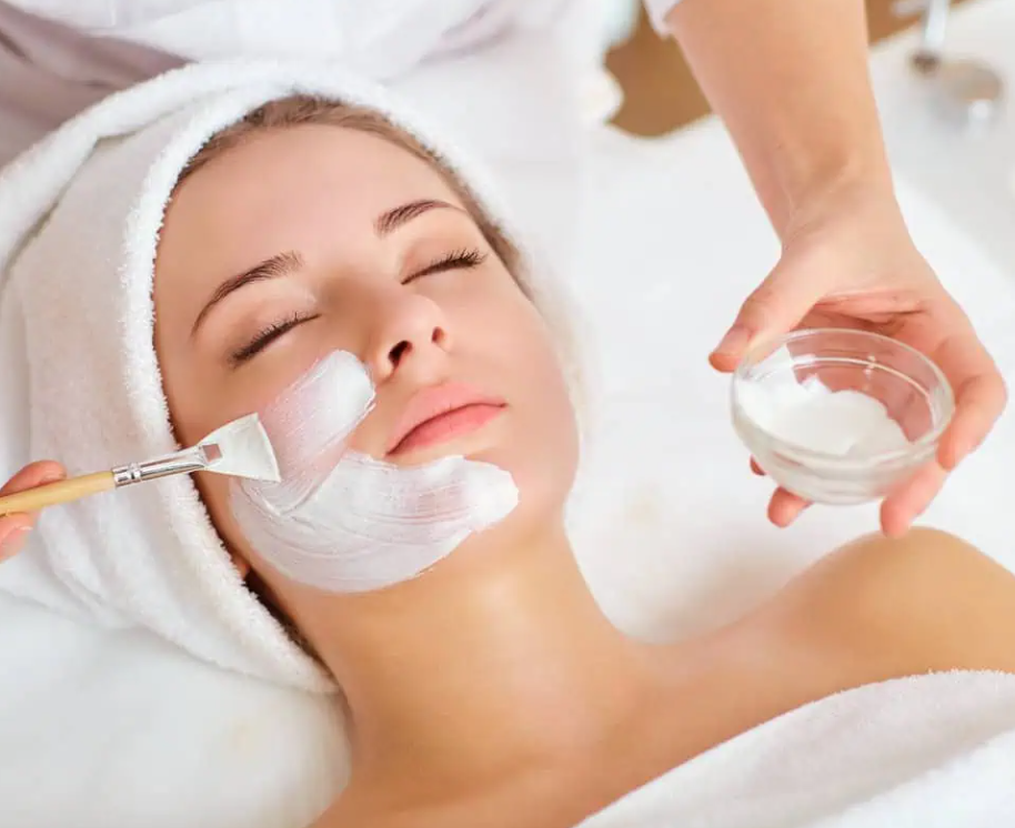 A woman is receiving a facial treatment, with her eyes closed, lying on a spa bed with a towel wrapped around her head. An aesthetician is applying a white facial mask using a brush and holding a bowl of the mask.