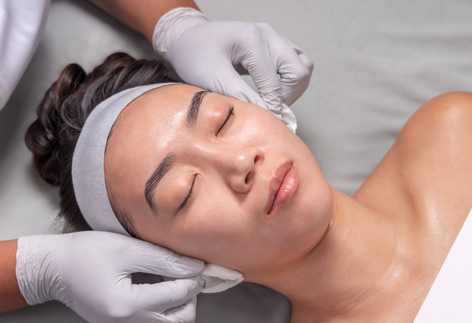 A woman receiving a facial treatment from an esthetician wearing white gloves.
