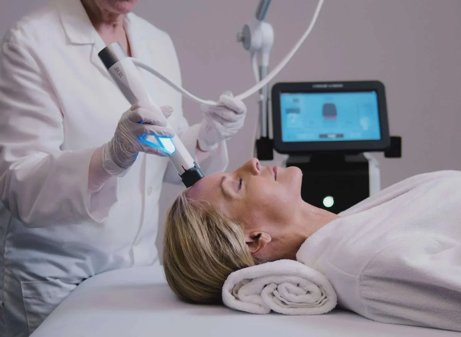 A woman lying on a treatment bed with a rolled towel under her head, receiving a facial procedure using a handheld device in a medical setting with a monitor in the background.