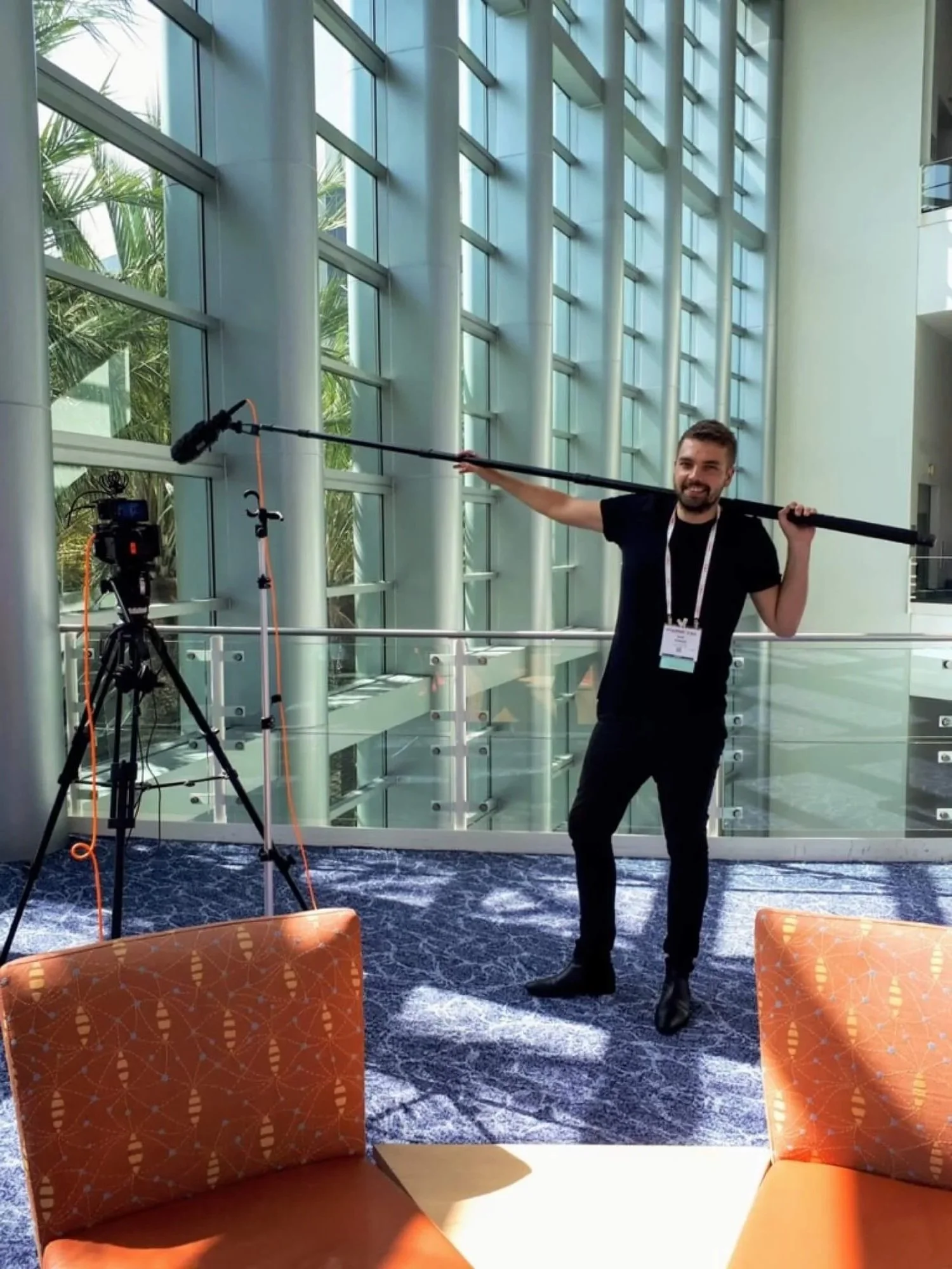 Man standing inside a building near large glass windows, smiling, holding a boom microphone over his shoulder, with a camera set up on a tripod nearby