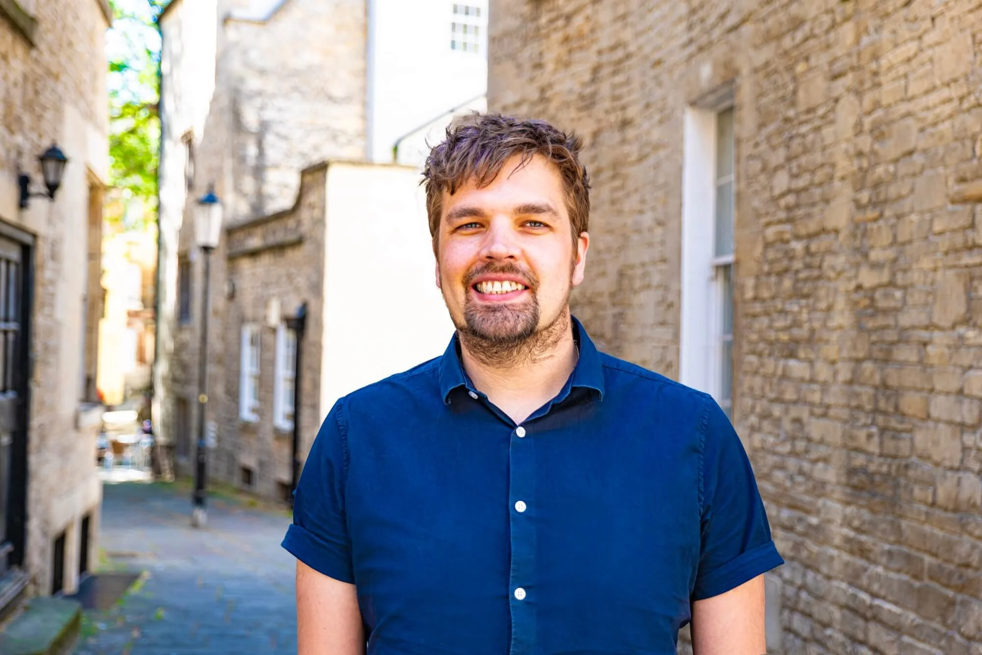 A smiling man with brown hair and a beard wearing a dark blue button-up shirt, standing outdoors between brick buildings with windows, lanterns, and a narrow paved alley in the background