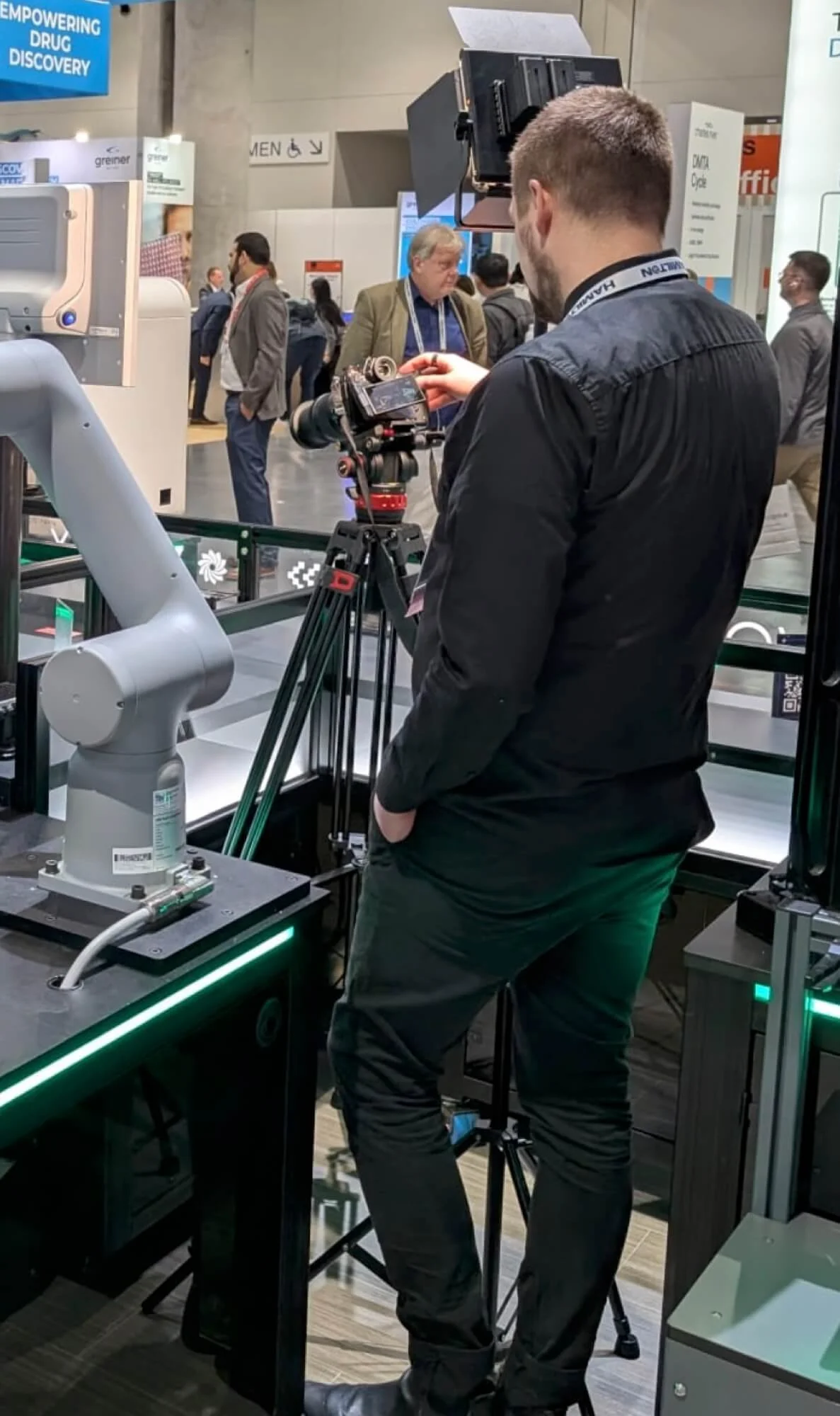 A man operating a camera on a tripod at a technology exhibition, with several people walking and talking in the background.