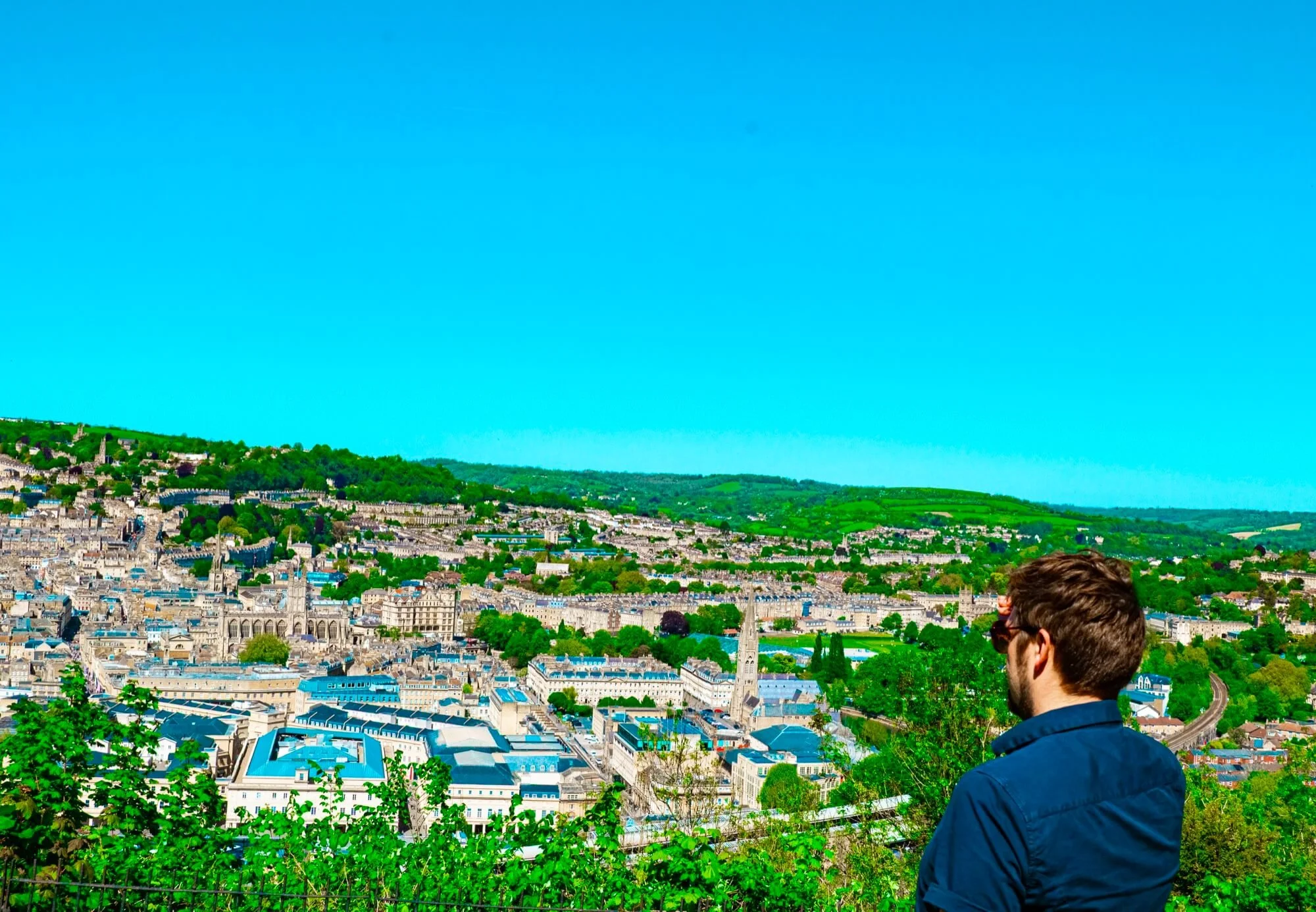 A man with brown hair and a beard, wearing a dark shape, gazes over a cityscape of Bath with historic buildings, green hills, and a bright blue sky