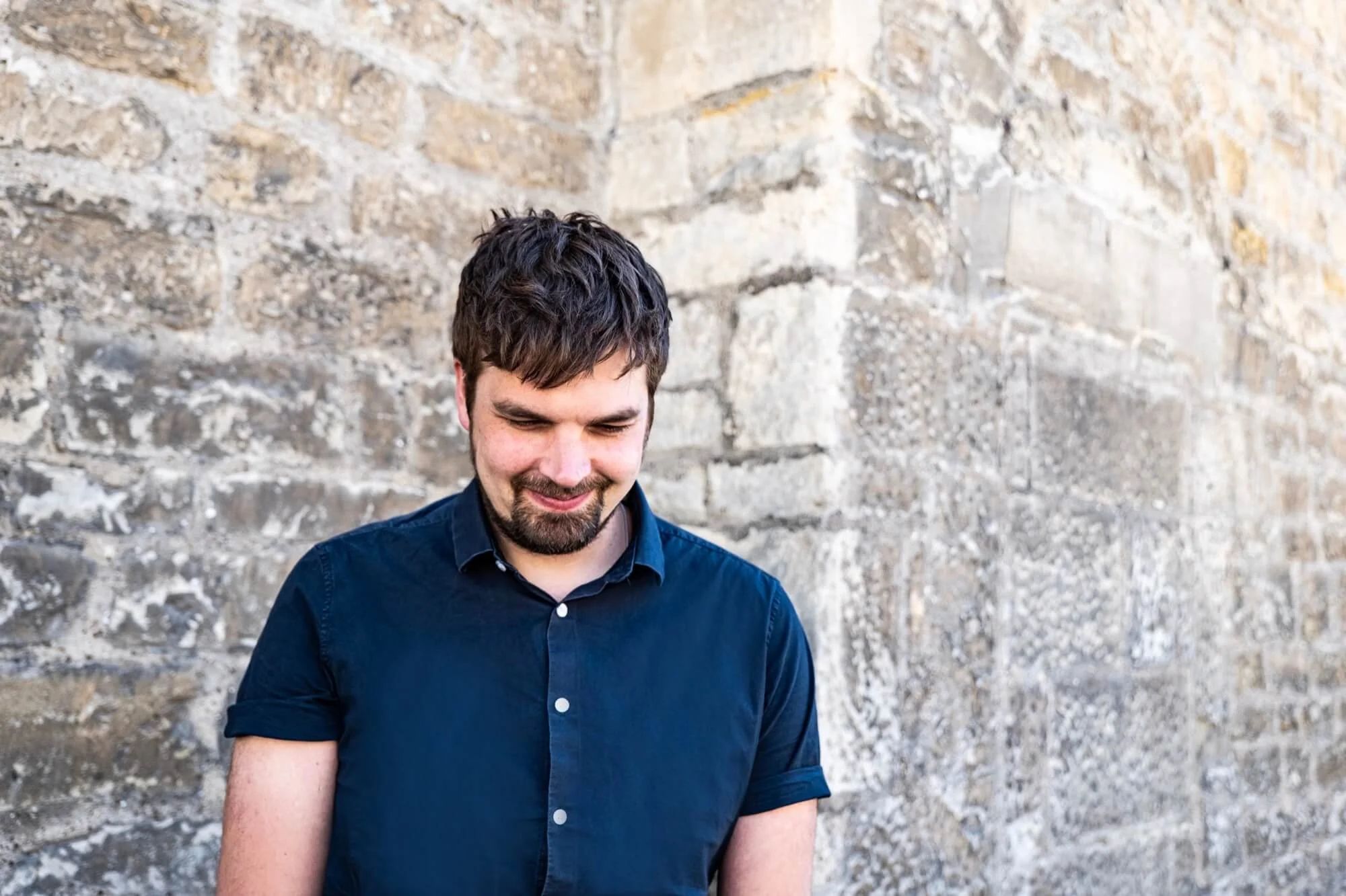 A  man with dark hair and a beard, smiling slightly and looking down, standing in front of a Bath stone wall.
