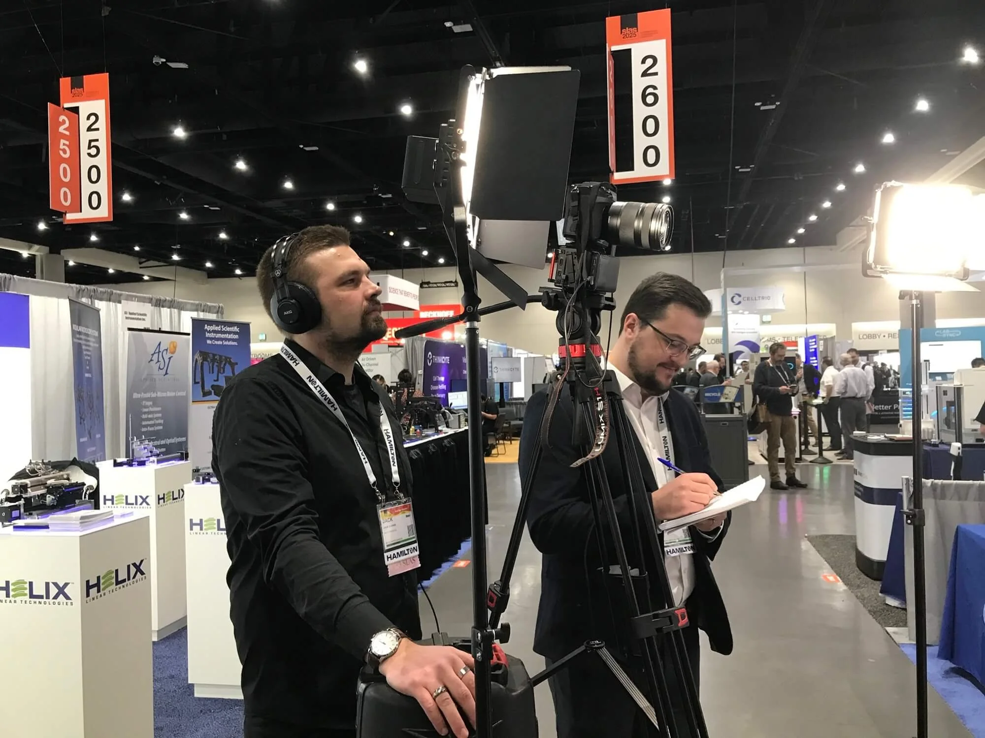 Two men operating a camera on a tripod at a conference, with booths and banners visible in the background