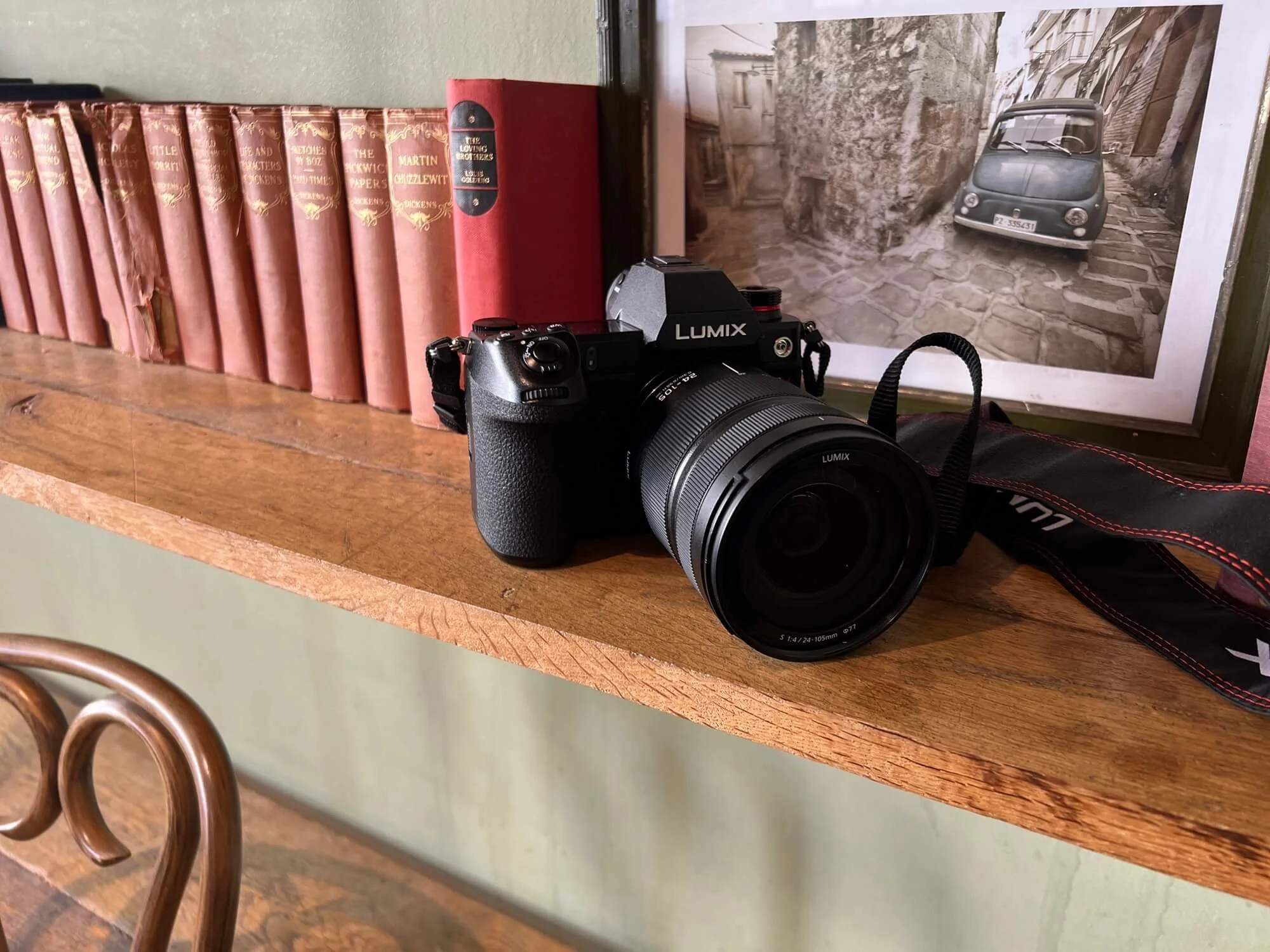 A black Lumix camera with a zoom lens resting on a wooden shelf, with old hardback books beside it
