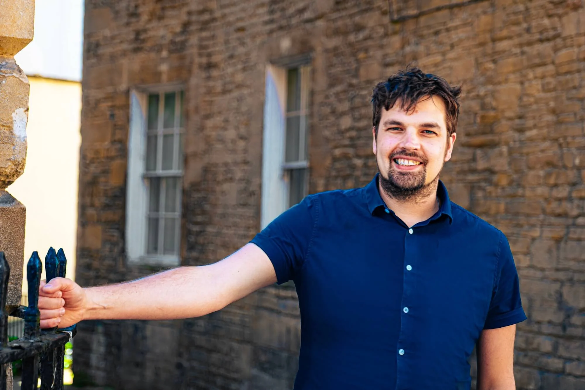 A smiling man with short dark hair and a beard, wearing a blue short-sleeved shirt, standing outdoors near a brick building, holding onto a black metal fence.