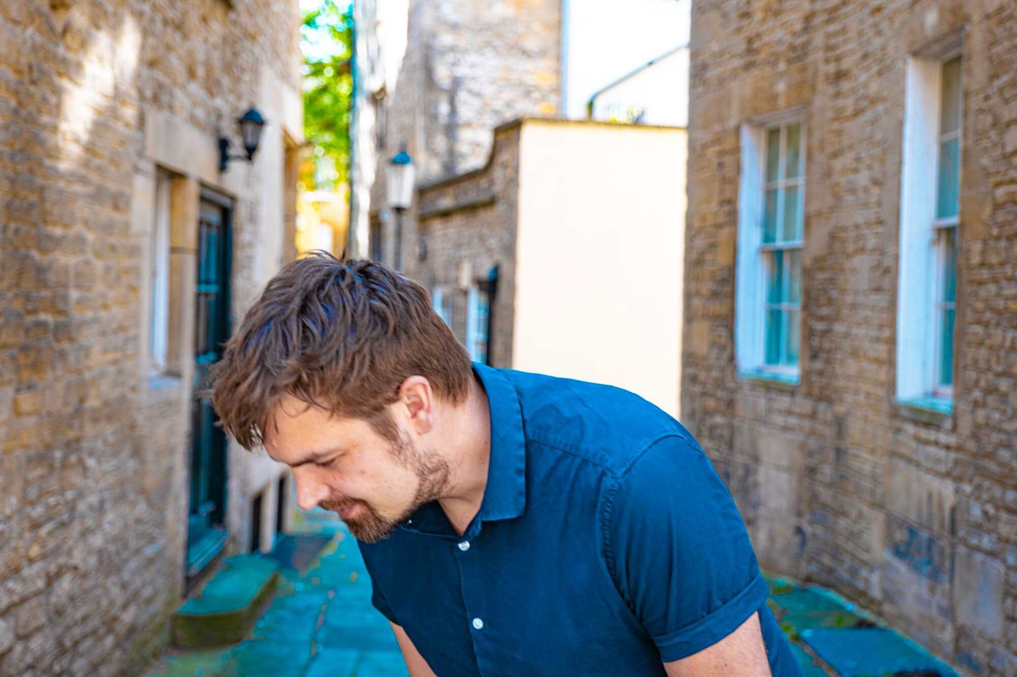 A man with brown hair and a short beard leaning forward and looking down, wearing a blue shirt, standing in a narrow alleyway with brick buildings and windows on either side