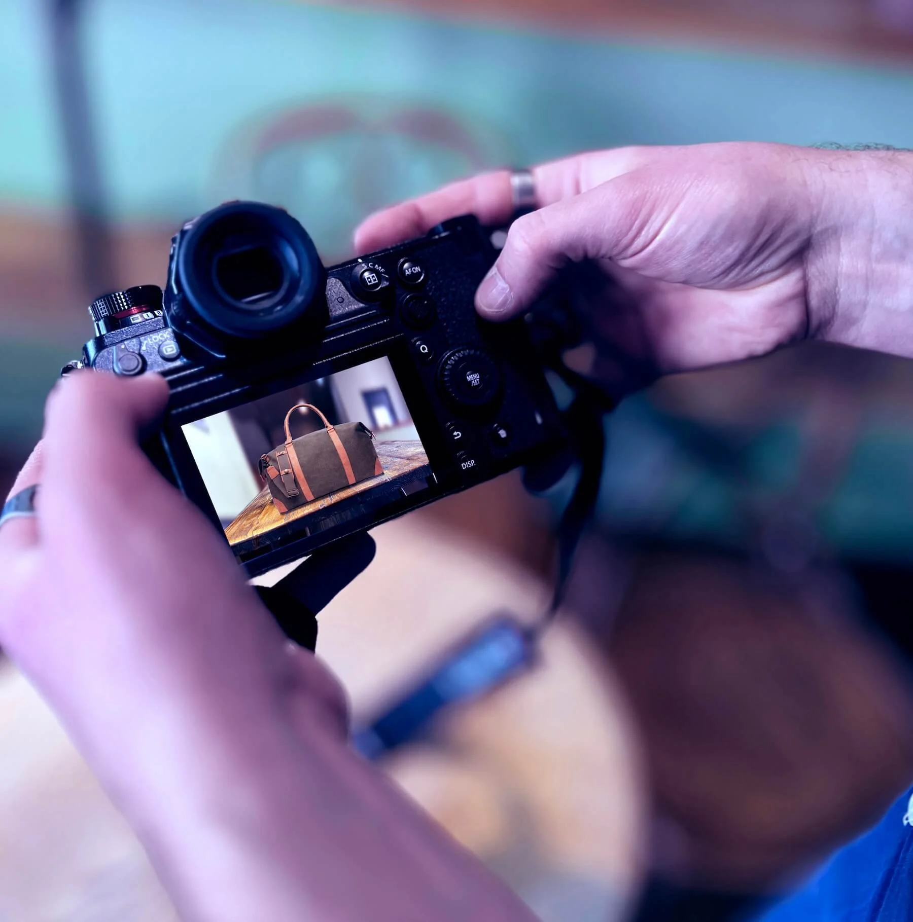 A person's hand holding a digital camera, capturing an image of a bag on a table. The camera's screen displays the bag in the frame.
