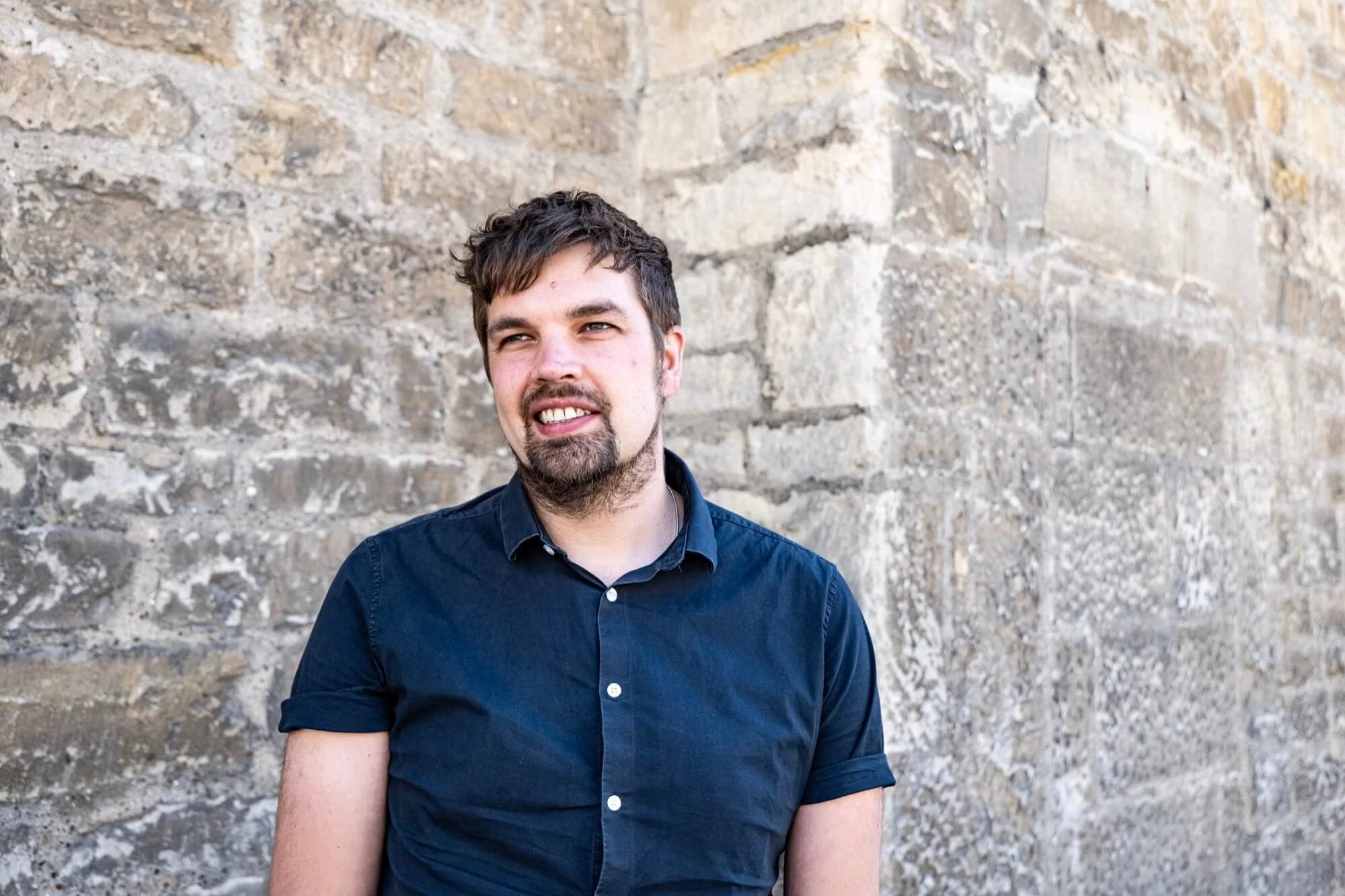 A  man with dark hair and a beard, wearing a dark blue button-up shirt, standing in front of a textured stone wall.