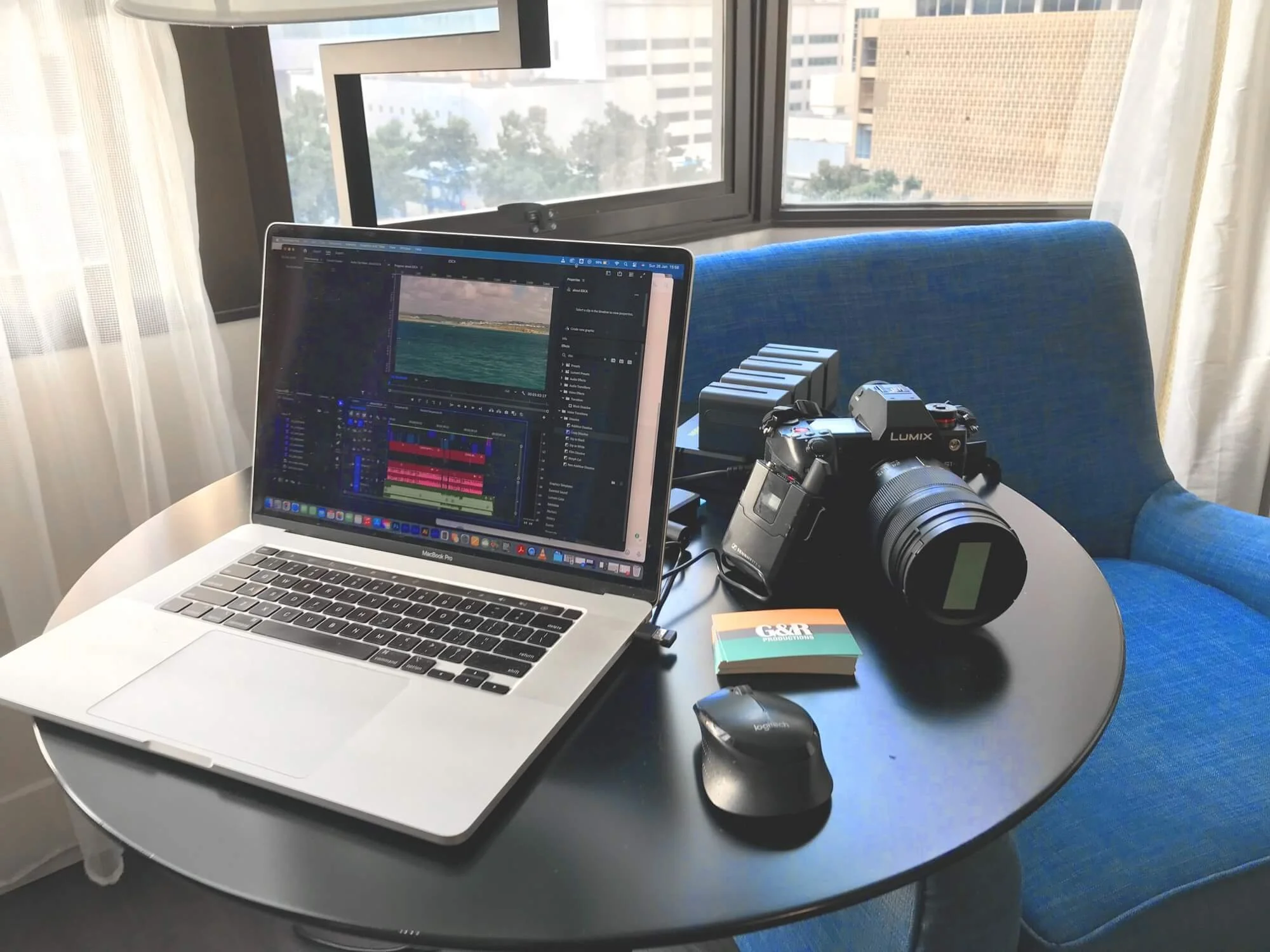 Indoor workspace with a laptop displaying video editing software, a camera, a mouse, and a stack of Goose and Racket business cards on a round black table