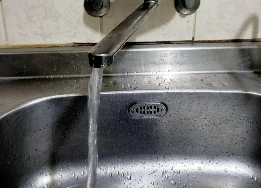 Running water pouring from a faucet into a stainless steel kitchen sink with water droplets on the sink surface.