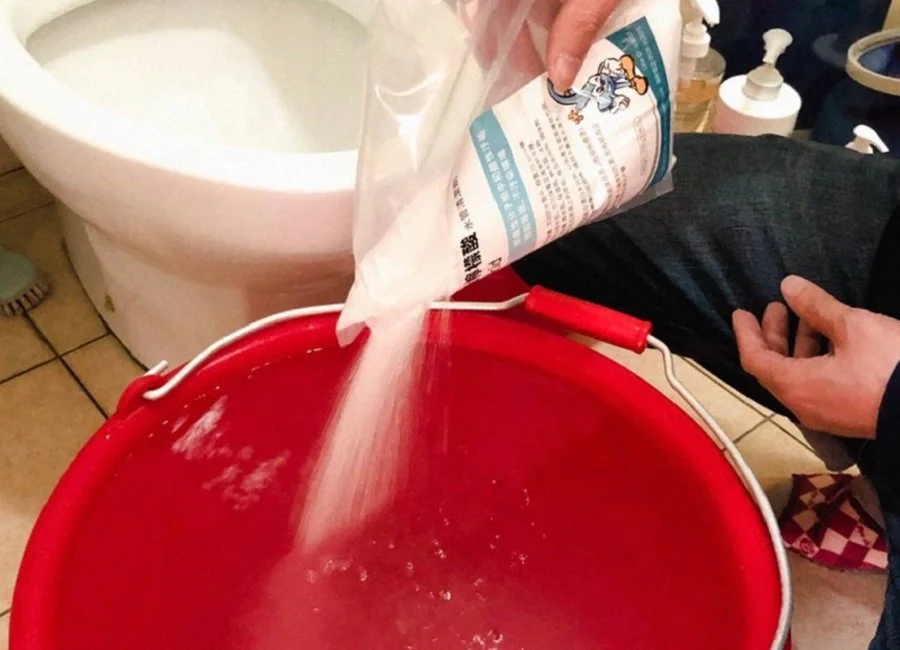 Person pouring powdered cleaning agent from a blue and white package into a red bucket in a bathroom.