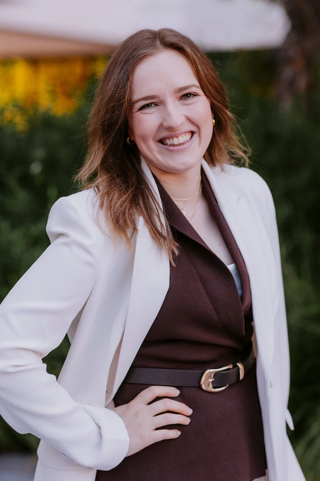 A smiling woman with shoulder-length brown hair, wearing a white blazer over a dark brown dress with a belt, outdoors with blurred green foliage in the background.