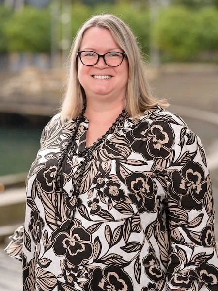 A woman with long blonde hair, glasses, and a floral patterned blouse standing outdoors near water.