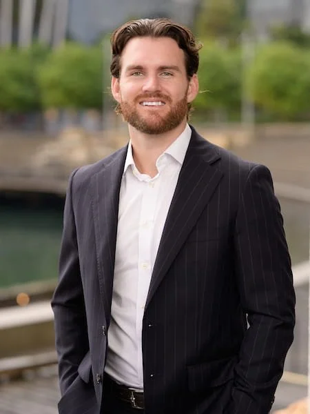 Man with brown hair and beard wearing a dark pinstripe suit and white shirt standing outdoors near a body of water, blurred cityscape background