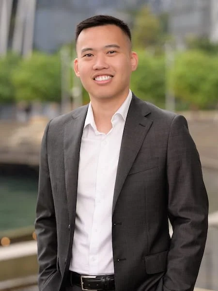 A man in a gray suit and white shirt smiling outdoors with blurred buildings and greenery in the background.