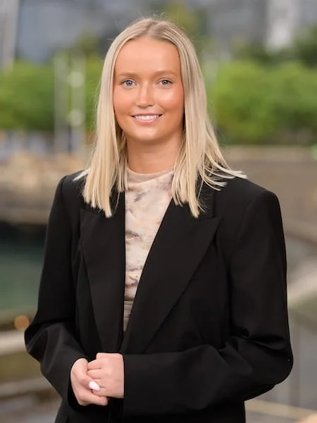 A young woman with blonde hair smiling, wearing a black blazer and a patterned top, standing outdoors with blurred greenery and water in the background.