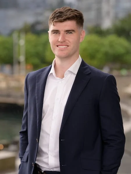 Young man in a suit smiling outdoors, with a blurred cityscape and water in the background.