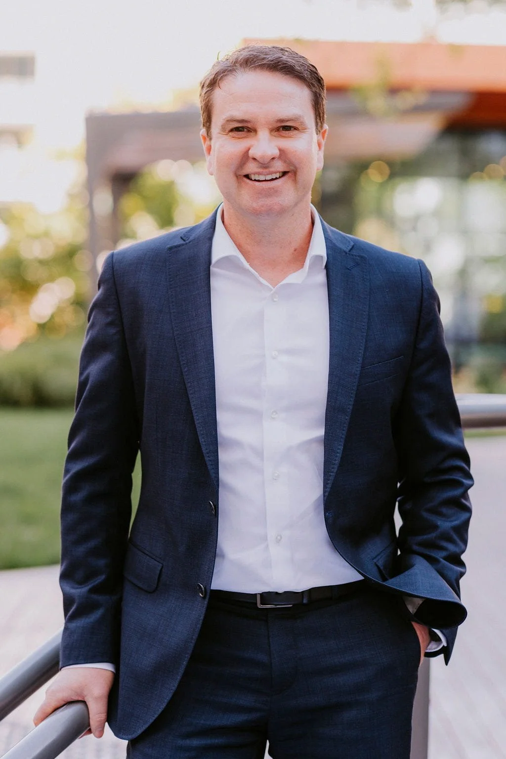 A man in a navy blue suit and white shirt standing outdoors, smiling, with his right hand resting on a railing.