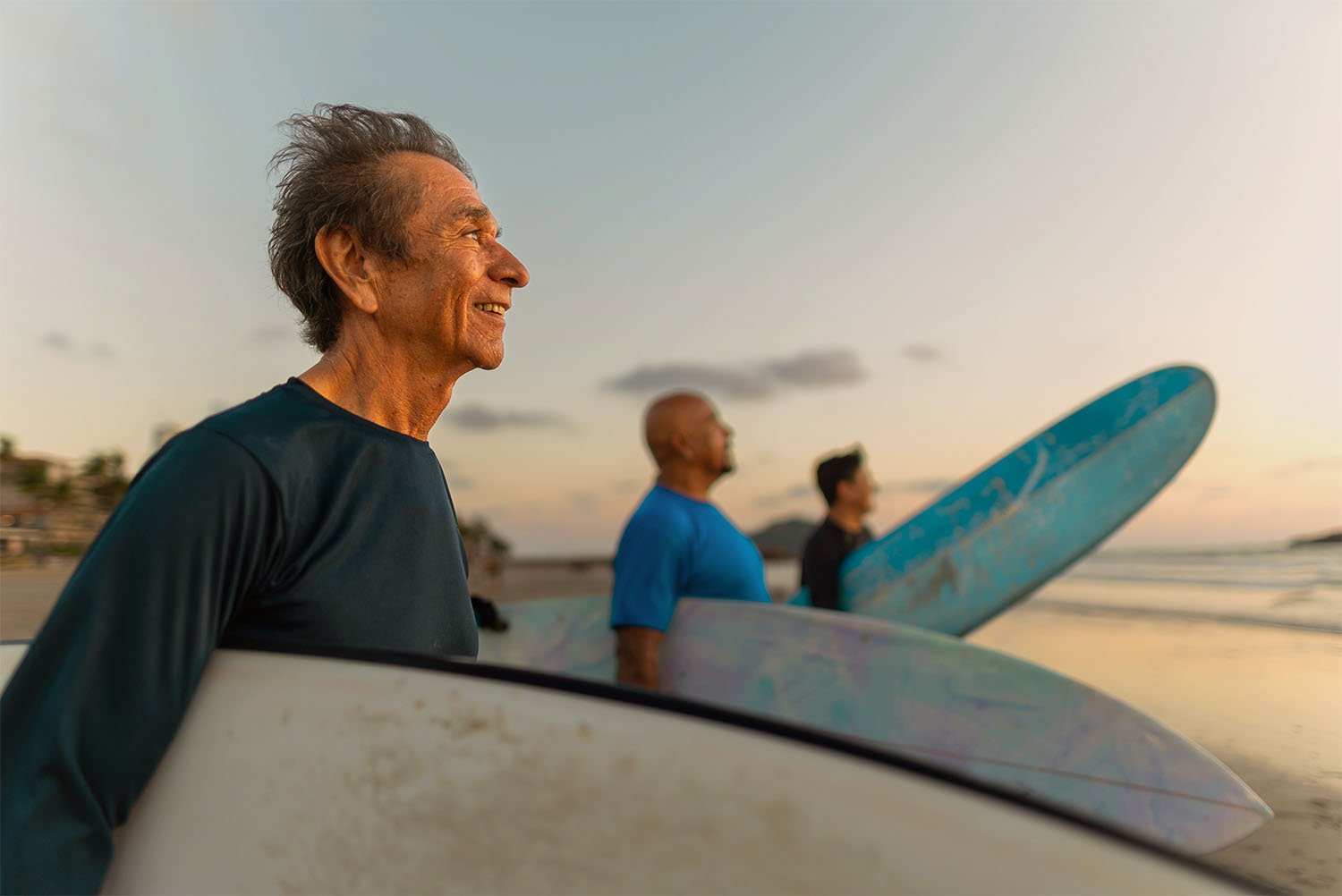 Three men standing on the beach with surfboards during sunset, smiling and looking out at the ocean.