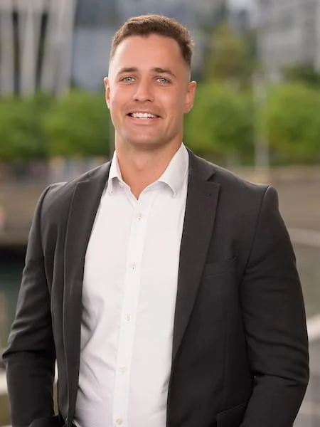 A man in a white shirt and black blazer standing outdoors with greenery and power lines in the background.