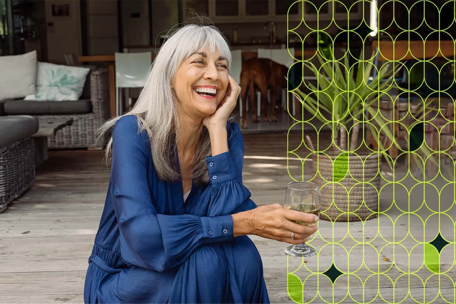 A joyful older woman with gray hair and a blue dress sitting on a wooden patio, holding a glass of white wine, smiling and relaxing indoors with a living room and dog in the background.