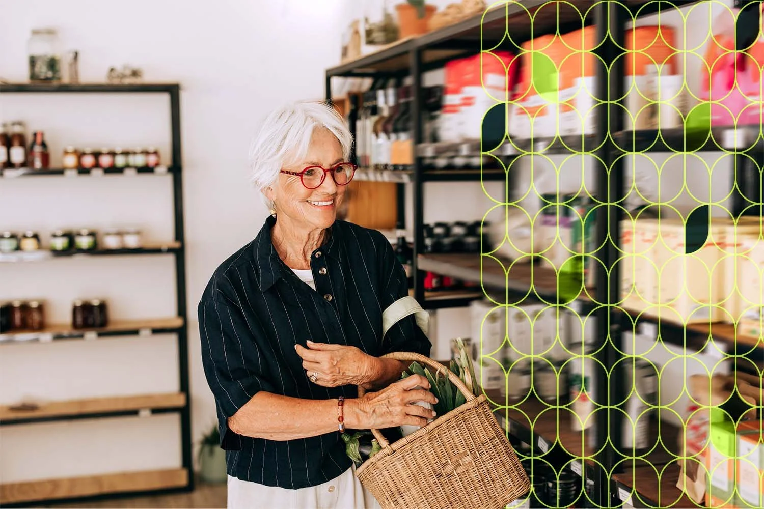 An elderly woman with white hair and red glasses shopping in a store, holding a wicker basket and smiling, surrounded by shelves with jars and containers.