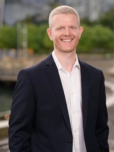 Portrait of a smiling man in a dark suit jacket and white shirt standing outdoors with a blurred city background.