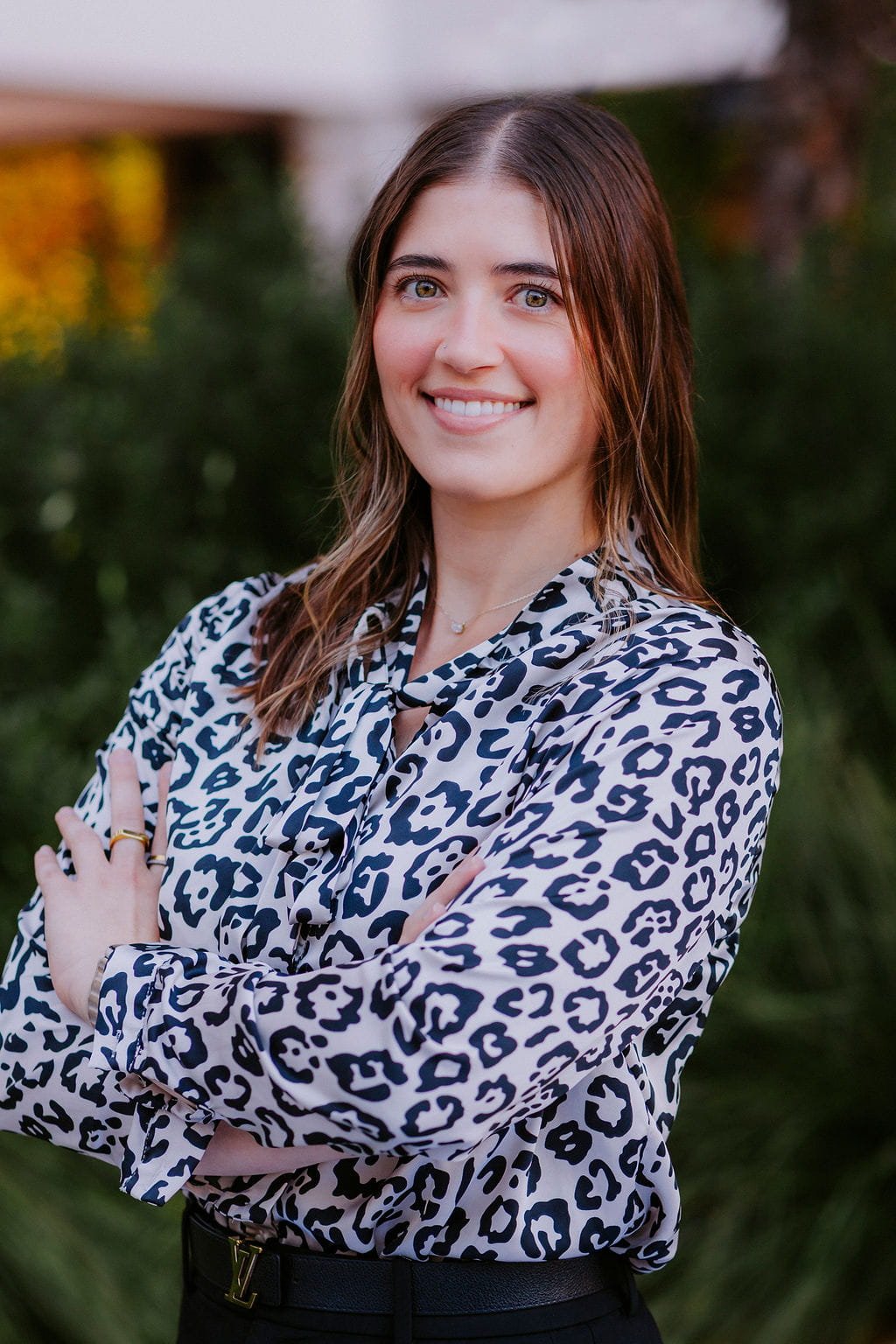 Young woman with light brown hair and green eyes, smiling, wearing a black and white animal print blouse and a small necklace, standing outdoors with greenery in the background.