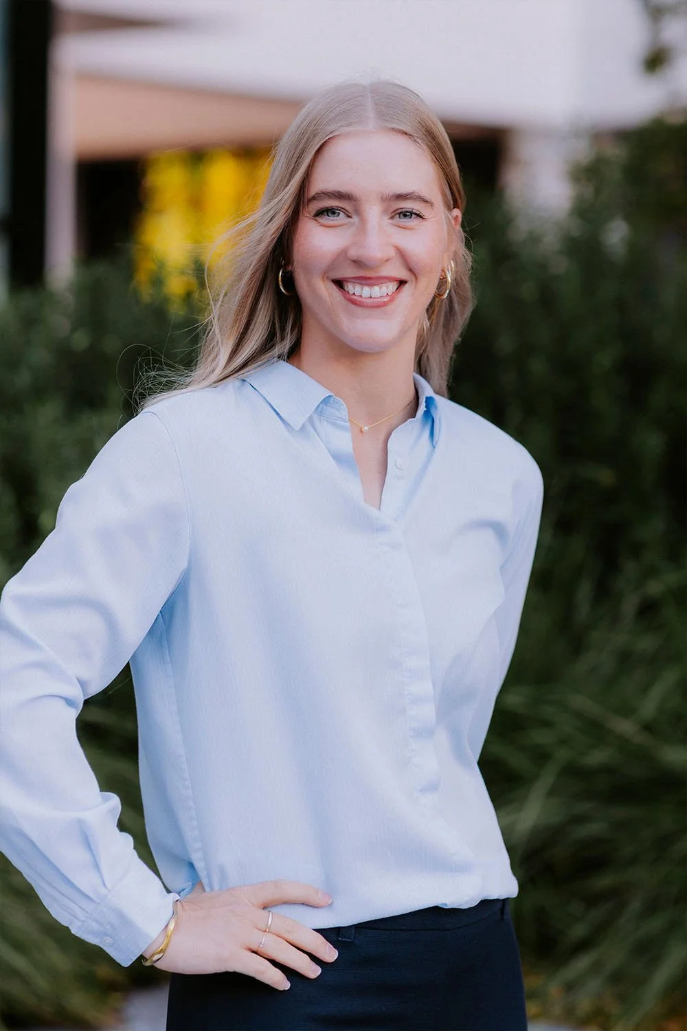A young woman with blonde hair, wearing a light blue button-up shirt, standing outdoors in front of greenery, smiling at the camera.