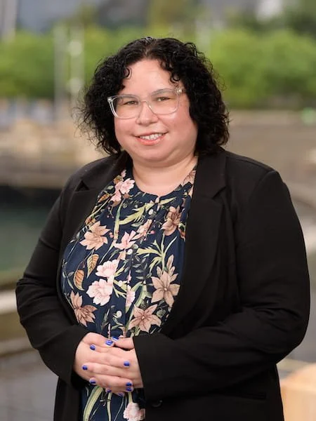 A woman with curly dark hair, wearing glasses, a black blazer, and a floral blouse, standing outdoors with a blurred background of trees and water.