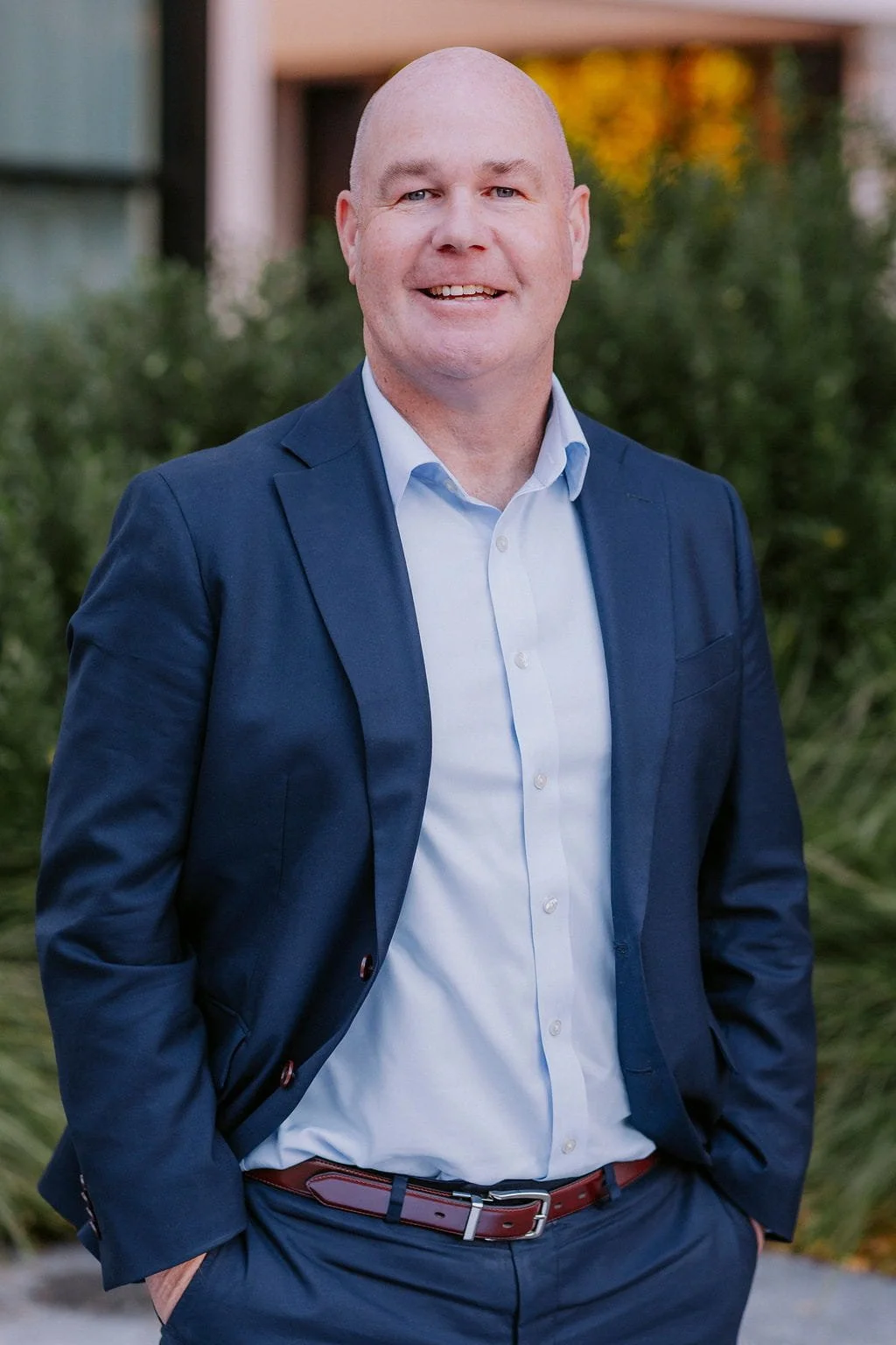 A bald man in a navy blazer and white shirt stands outdoors with his hands in his pockets, smiling. Background features green foliage and blurred structures, suggesting a professional photo.