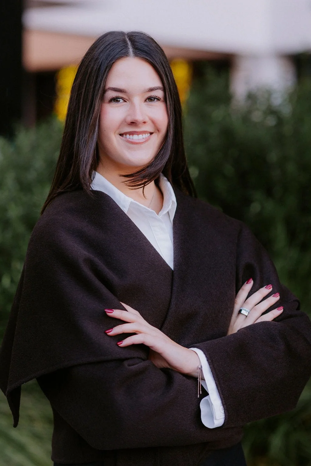A young woman with shoulder-length dark hair, smiling, wearing a dark academic gown over a white shirt, standing outdoors with greenery in the background.