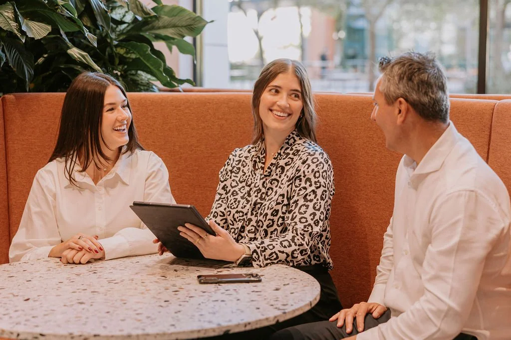 Up Wealth financial planner South Coast NSW staff sitting at a round table, engaged in conversation in pre-retirement financial strategy and smiling, in a cafe or restaurant with plants and large windows in the background.