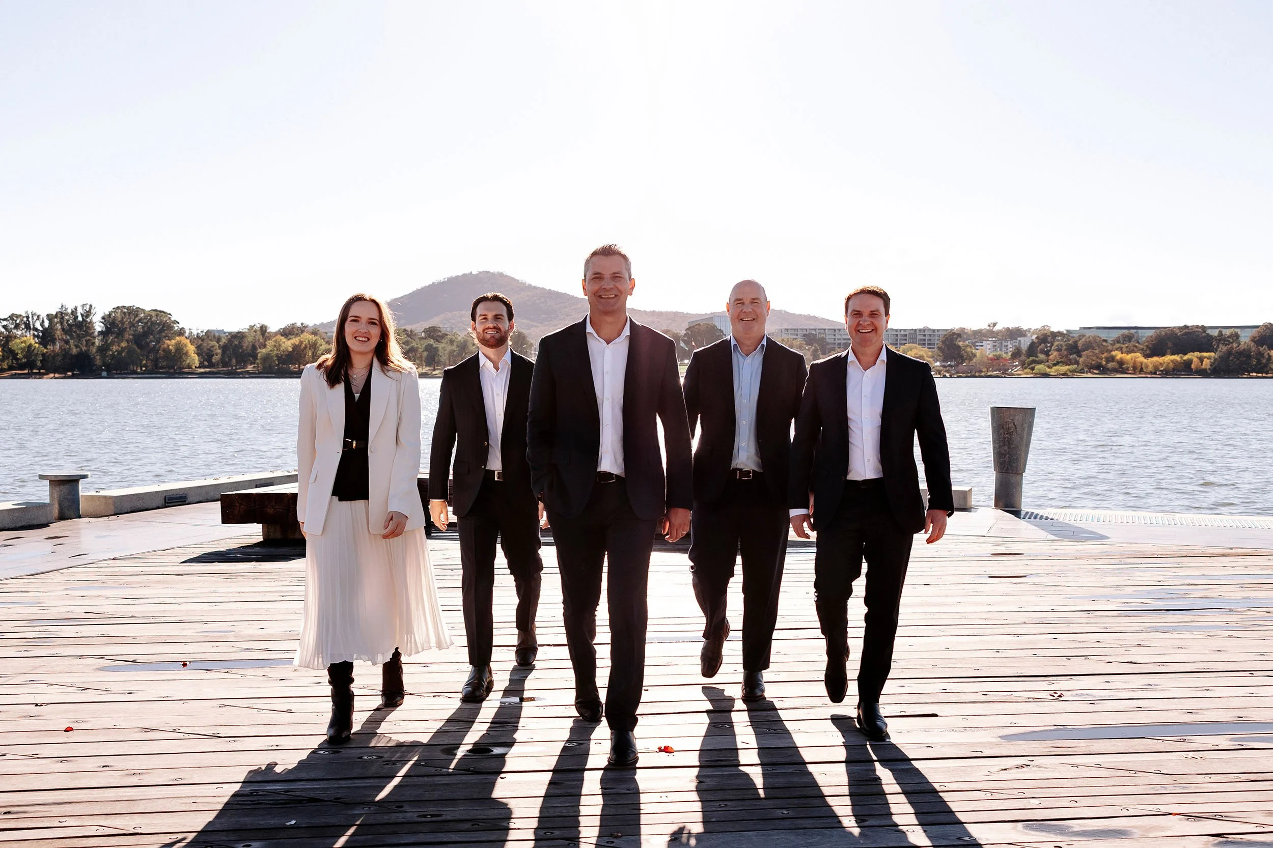 Up Wealth financial planner South Coast NSW professionally dressed walking along a wooden dock by a lake, with trees and buildings in the background and mountains in the distance, under a clear sky.