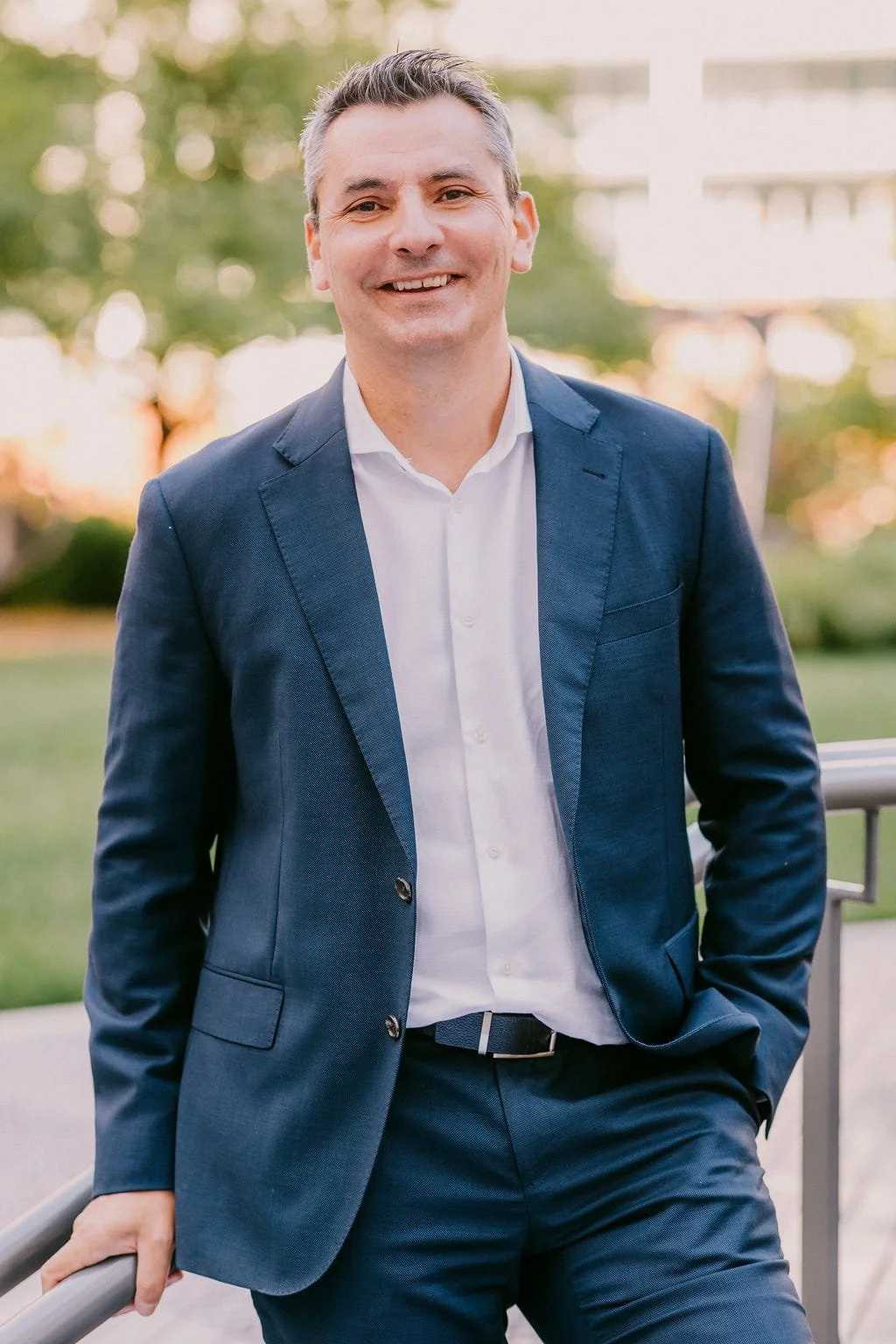A smiling man in a blue suit and white shirt standing outdoors in a park during sunset.