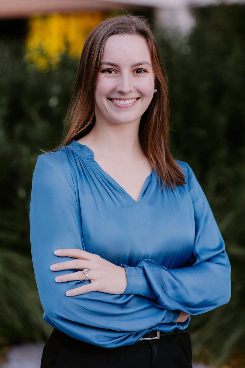 A smiling woman with straight, shoulder-length brown hair wearing a blue blouse with long sleeves, standing outdoors with greenery in the background.