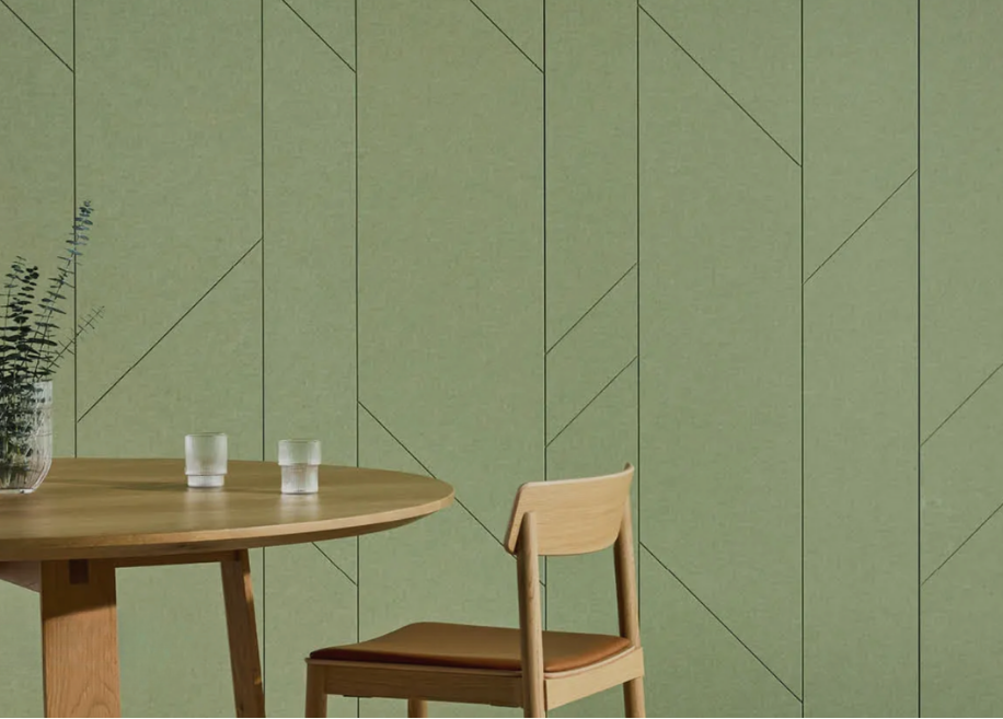 A minimalist dining area with a round wooden table, a single wooden chair, a potted plant, and two glasses of water against a green wall with geometric lines.