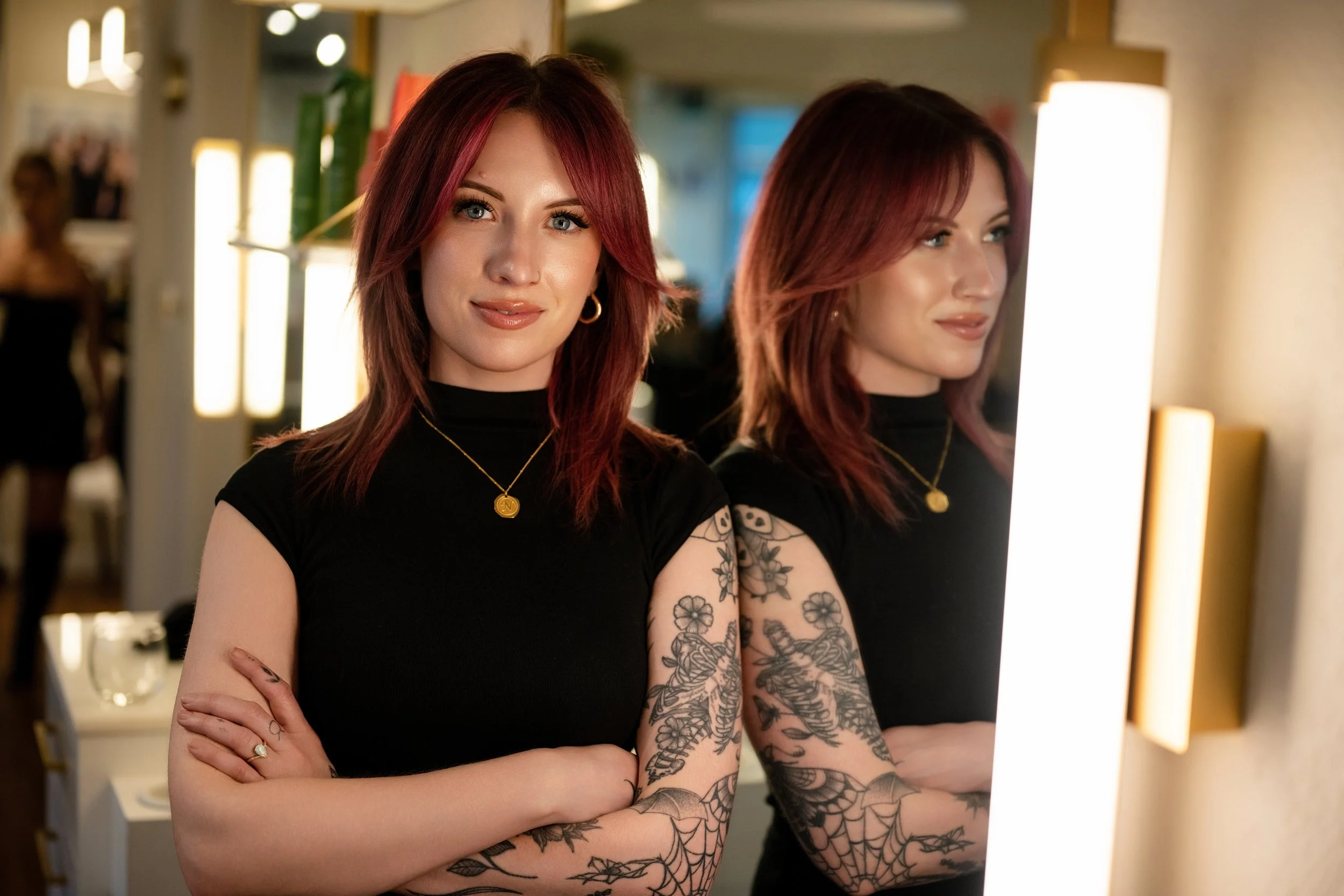 Two young women with red hair and tattoos standing side by side in front of a mirror in a makeup or hair salon, both looking at themselves with slight smiles.