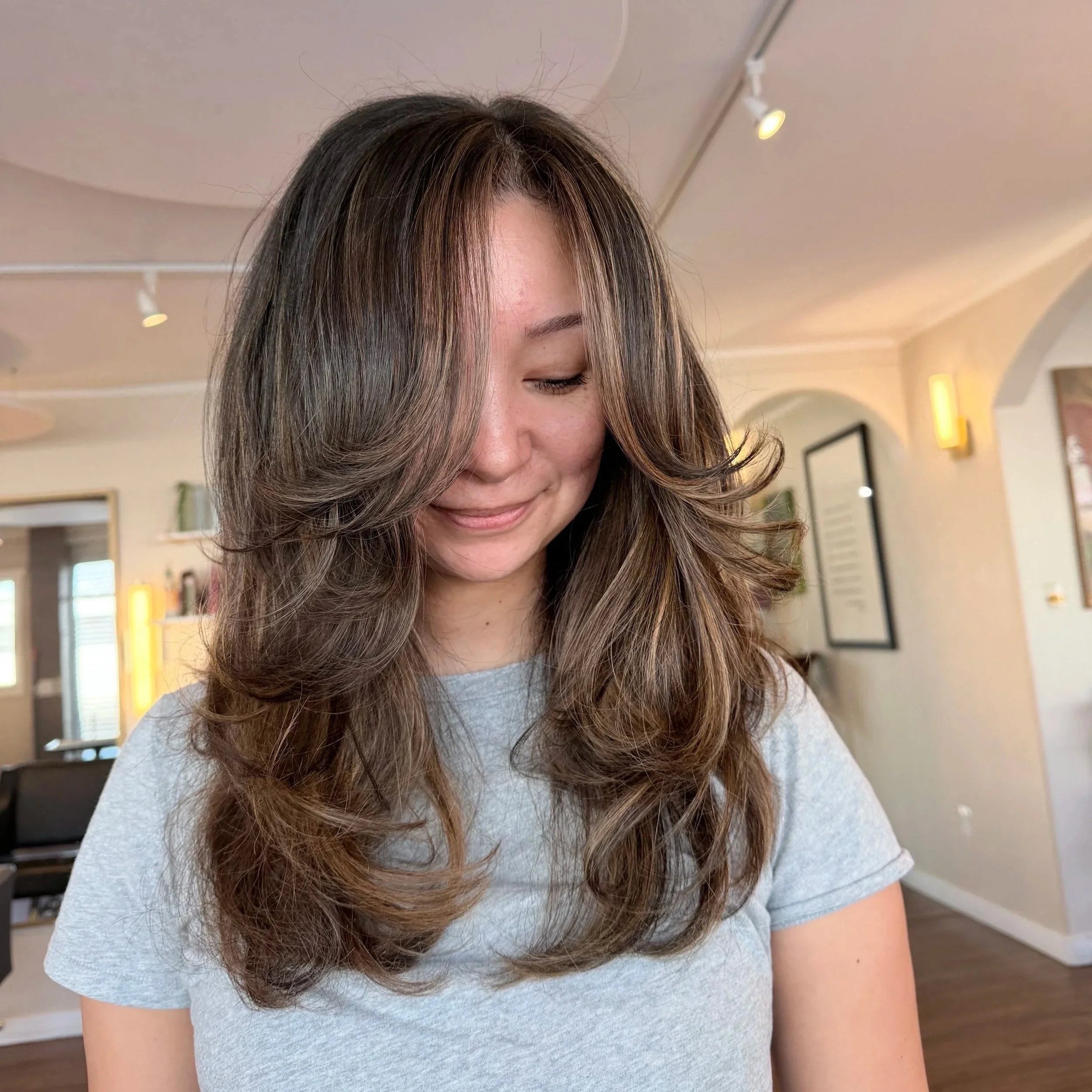 Close-up of a woman with wavy brown hair wearing a light gray shirt inside a modern room with warm lighting and framed pictures on the wall.