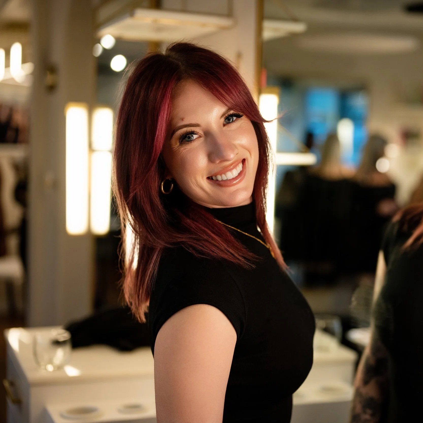 A woman with shoulder-length red hair and blue eyes smiling in a warmly lit indoor setting, wearing a black top and gold earrings.