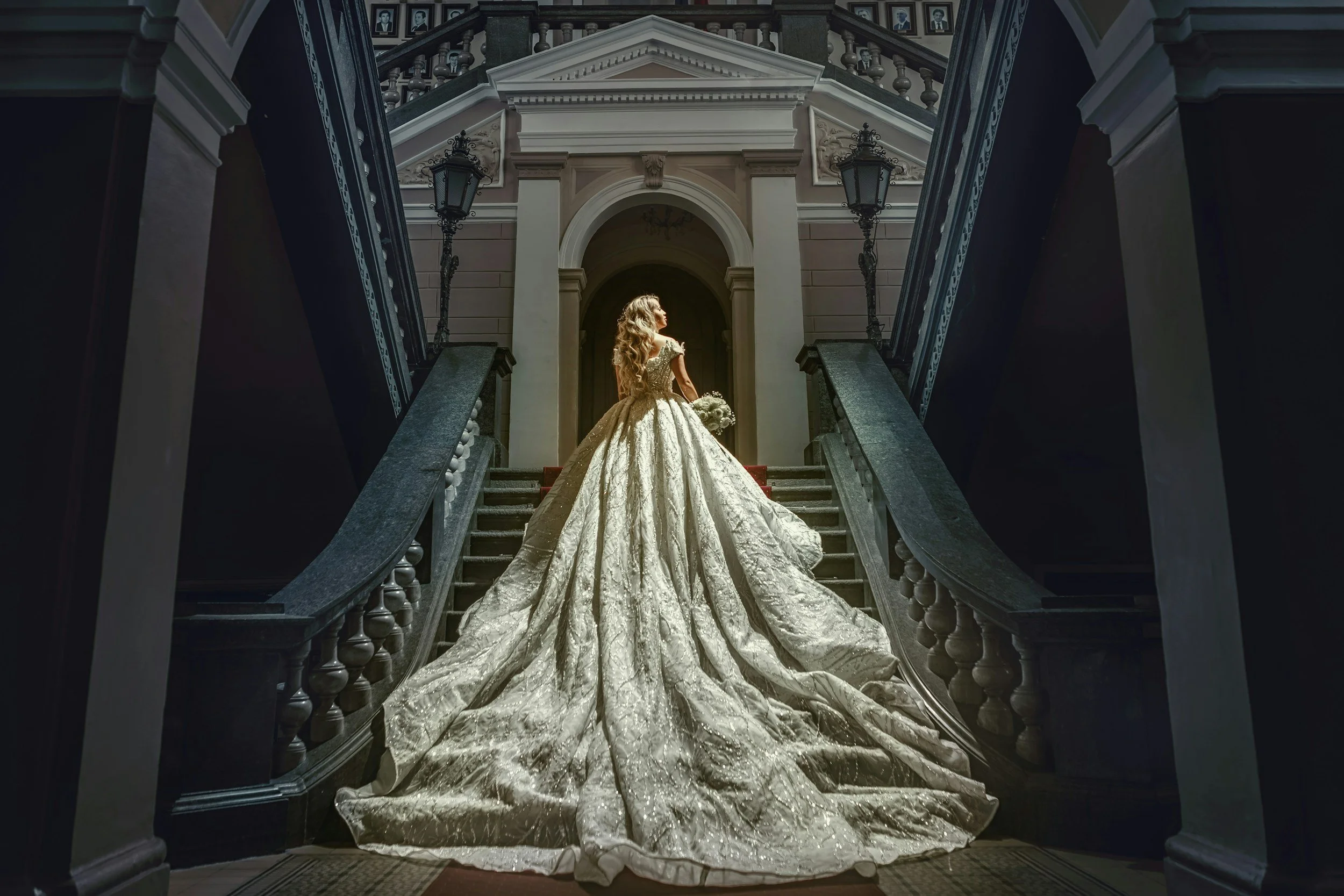 A woman in an ornate wedding gown with a long train stands on the stairs of a grand historic building, holding a bouquet and illuminated by sunlight.
