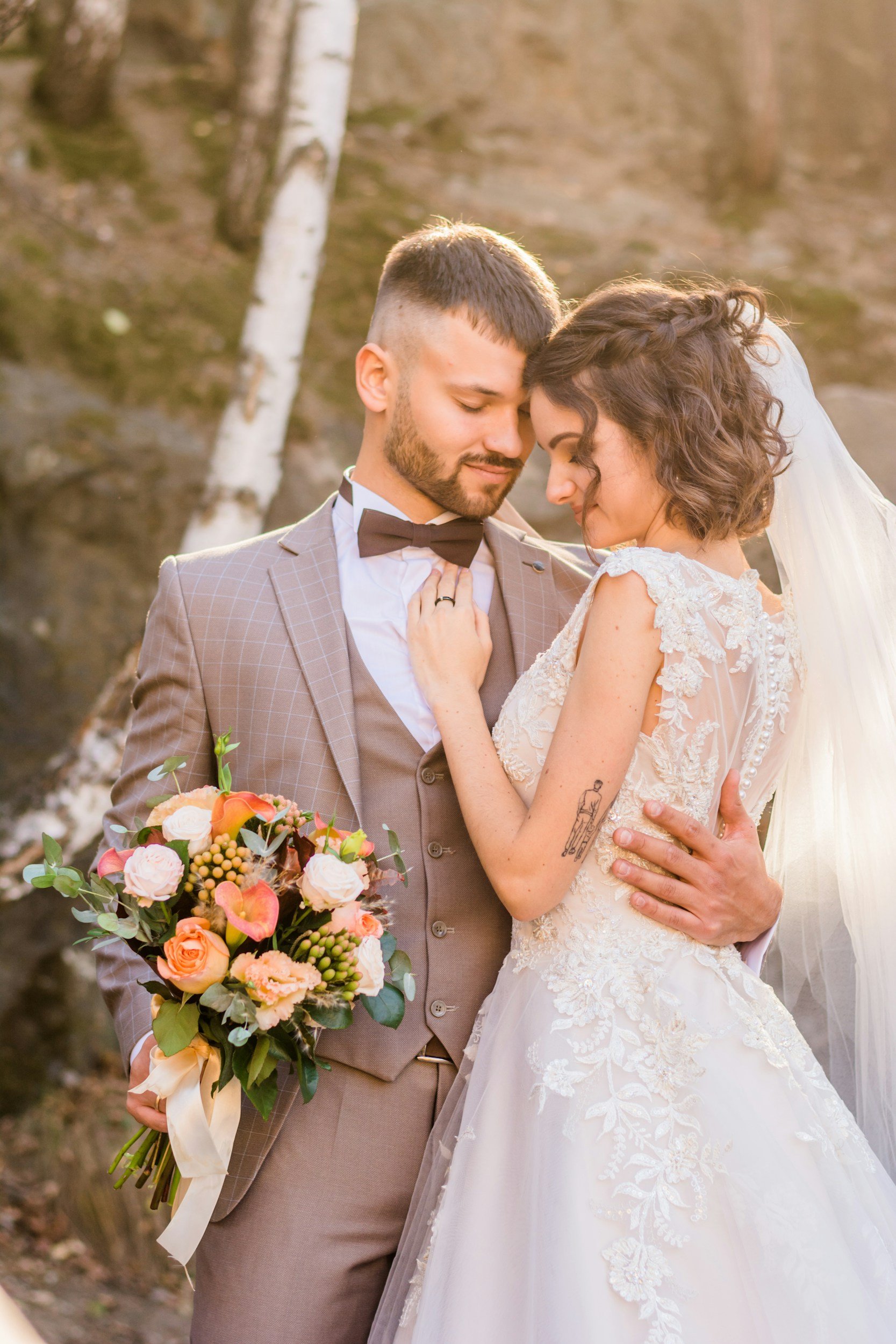 A bride and groom share a romantic moment outdoors, with the bride holding a flower bouquet, and trees in the background during sunset.
