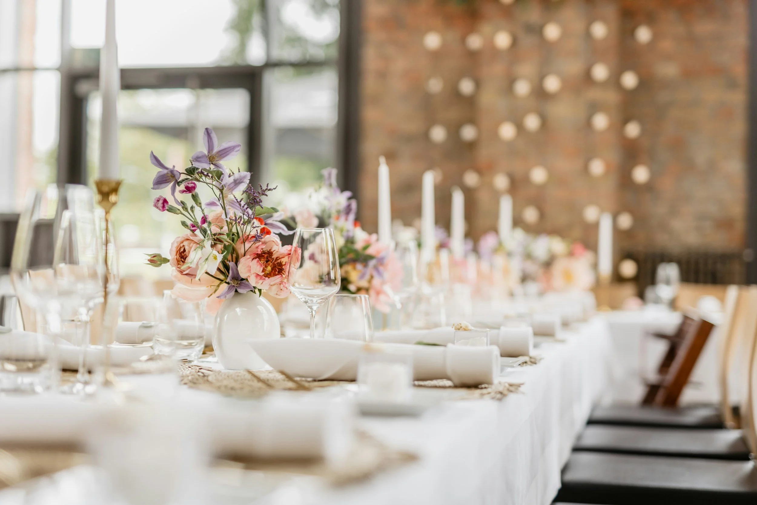 A table with a floral centerpiece of pink and purple flowers in a white vase, set with wine glasses, candles, and white plates in a bright, industrial-style event space with brick walls and large windows.