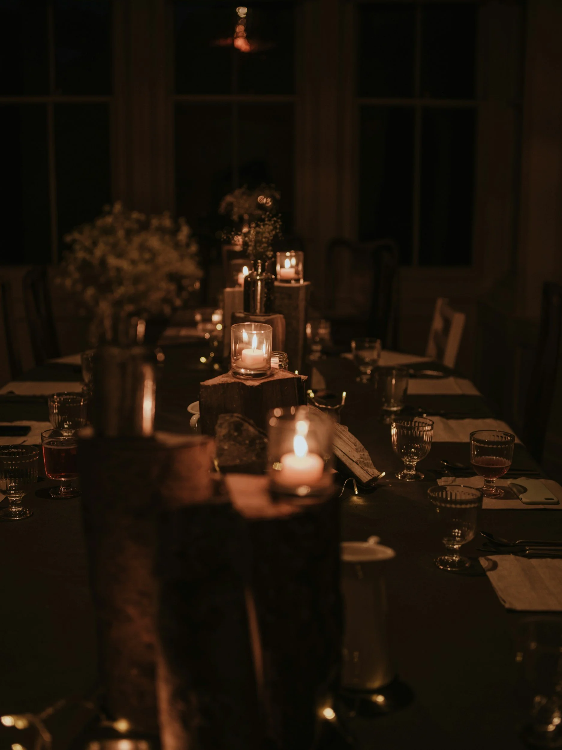 A darkly lit dining room decorated for an event with candlelit centerpieces, flowers, and tableware, viewed from across the table at night.