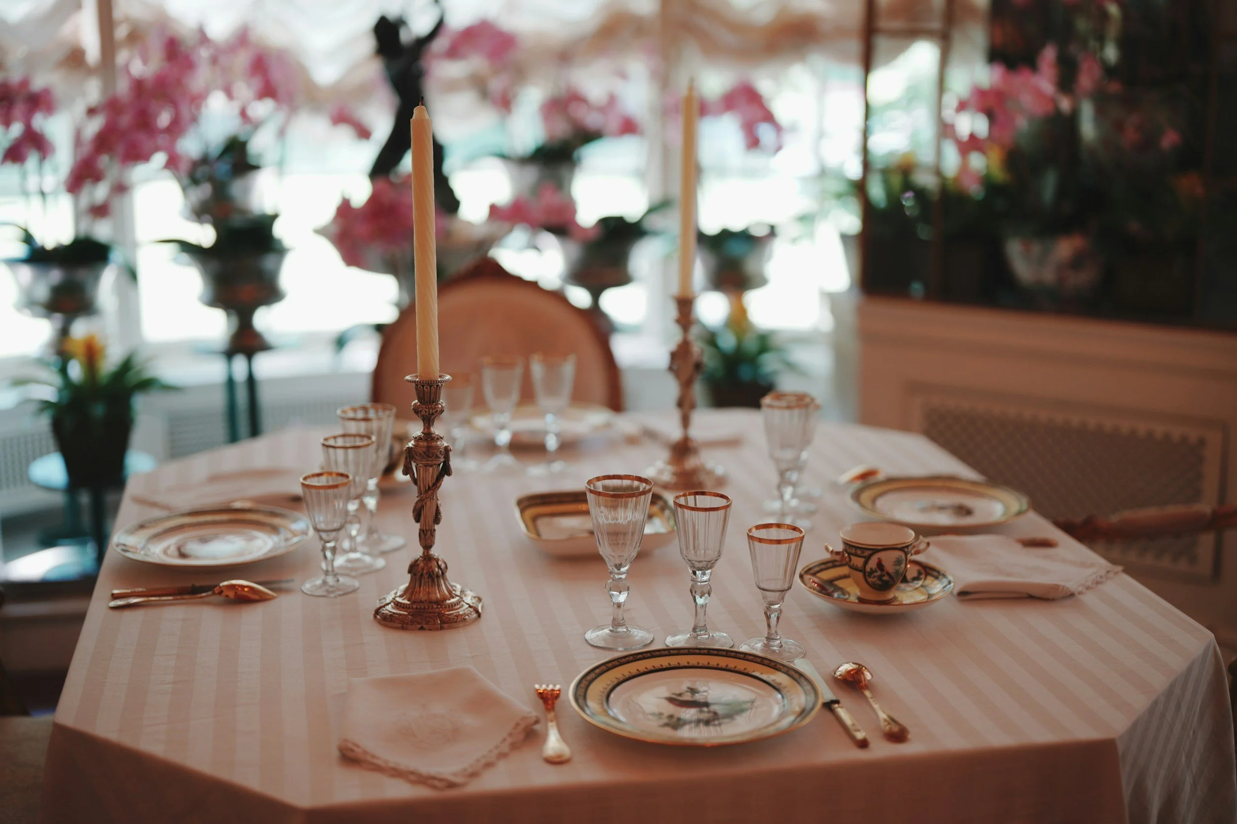 Elegant dining table set with fine china, glassware, and tall candles in a well-lit room decorated with pink flowers
