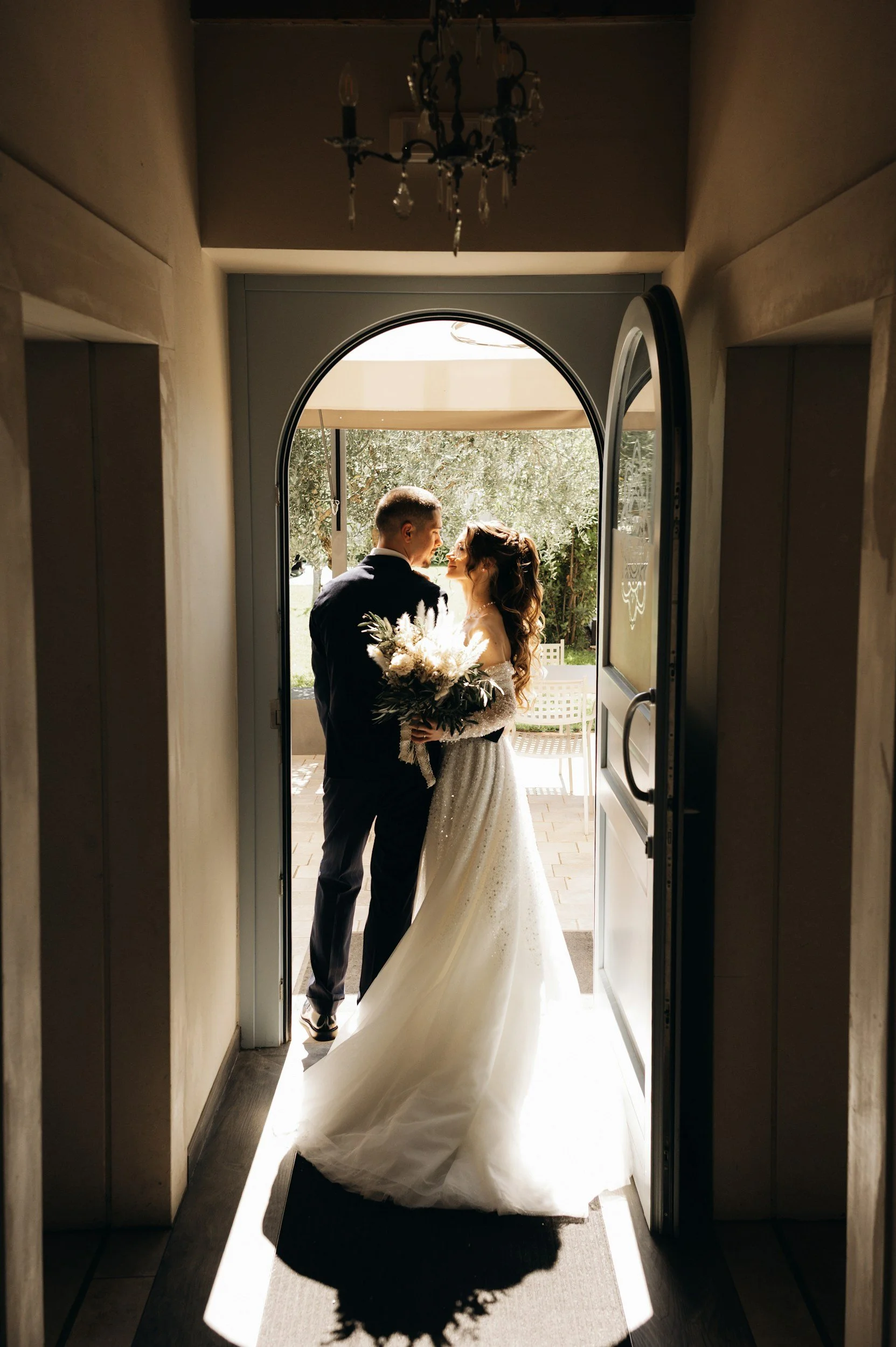 Bride and groom standing at the entrance of a building, holding hands and looking at each other, with sunlight casting shadows on the floor.