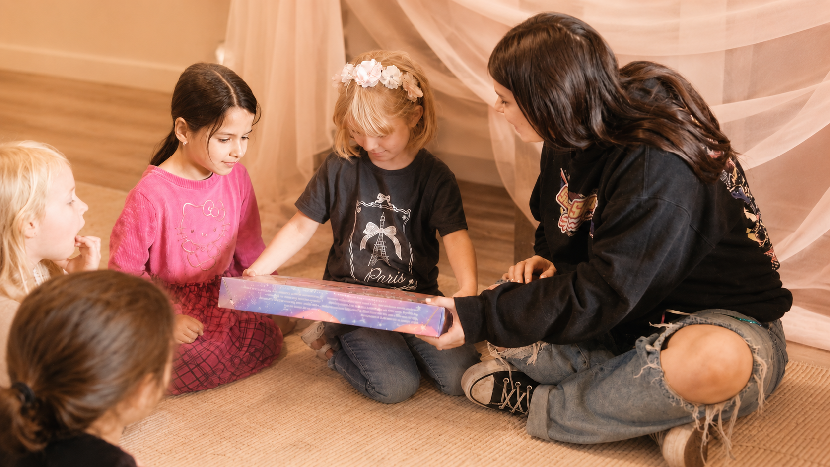 A woman and five young girls are sitting on a beige carpet, with the woman handing a colorful gift box to one of the girls. The girls are gathered around, with some looking at the gift and others looking at the woman, in a cozy indoor setting with soft curtains in the background.