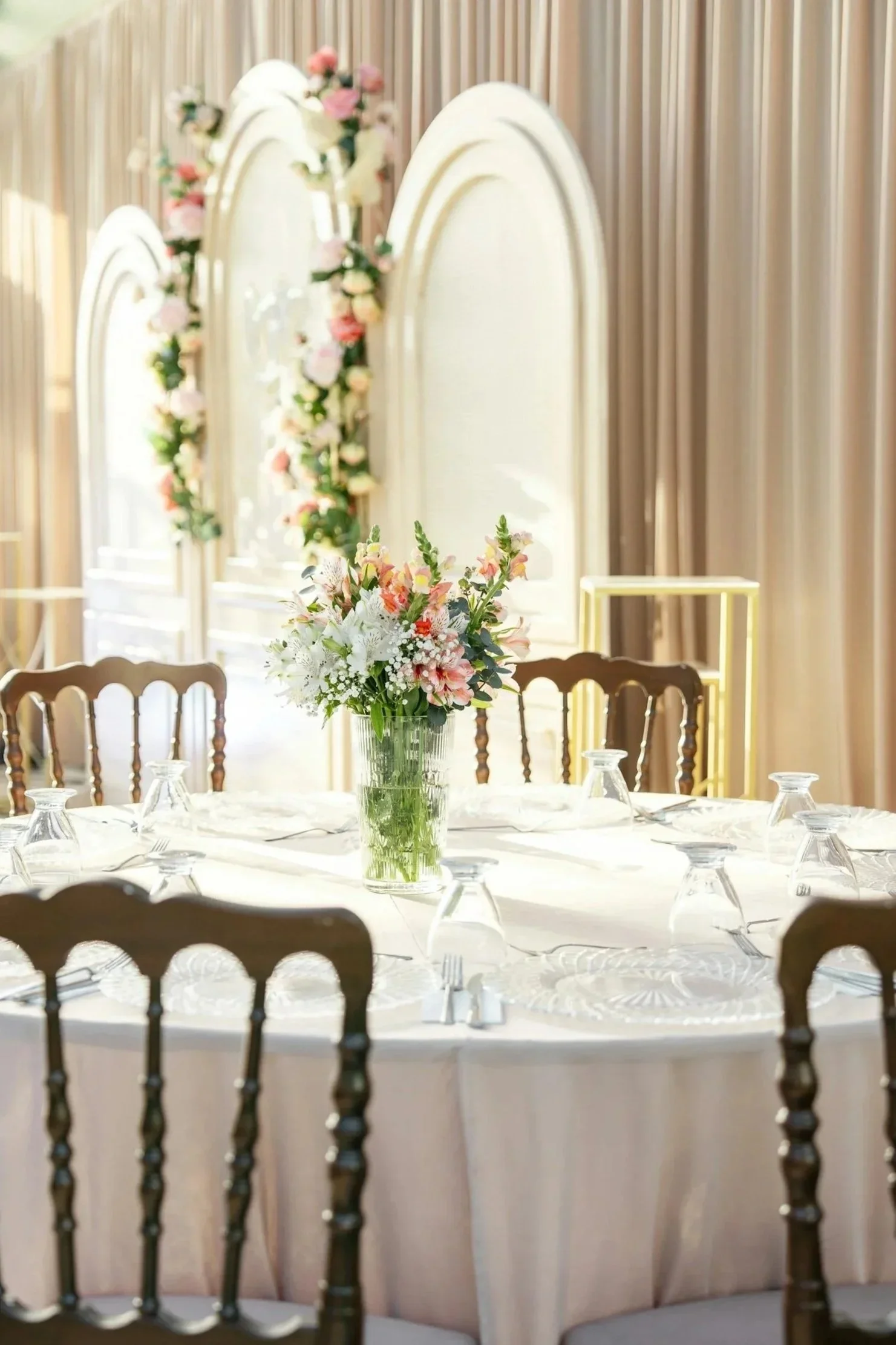 A round banquet table with a white tablecloth, set with glassware and a flower arrangement in a glass vase, with wooden chairs around it, in a room with beige curtains and three arched mirrors decorated with pink and white flowers.
